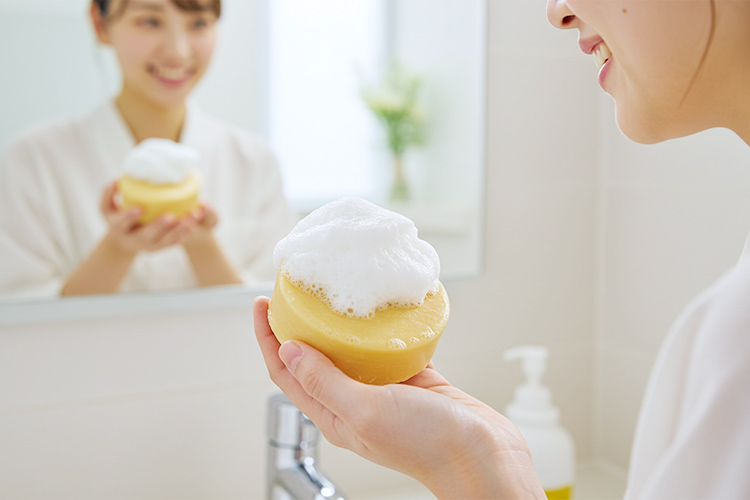 泡立つ丸い洗顔石鹸を手に持つ日本人女性の笑顔、バスルームの背景