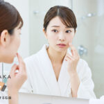 鏡を見ながら毛穴のつまりを気にしている日本人女性。背景は明るいバスルームで、洗面台と泡立てた洗顔料が映っている。