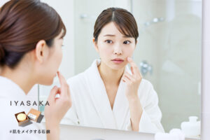鏡を見ながら毛穴のつまりを気にしている日本人女性。背景は明るいバスルームで、洗面台と泡立てた洗顔料が映っている。