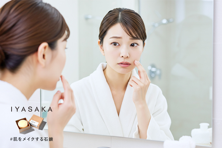 鏡を見ながら毛穴のつまりを気にしている日本人女性。背景は明るいバスルームで、洗面台と泡立てた洗顔料が映っている。