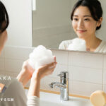 笑顔の女性が鏡の前で泡立てた洗顔石鹸の泡を手にのせ、肌をケアしているシーン